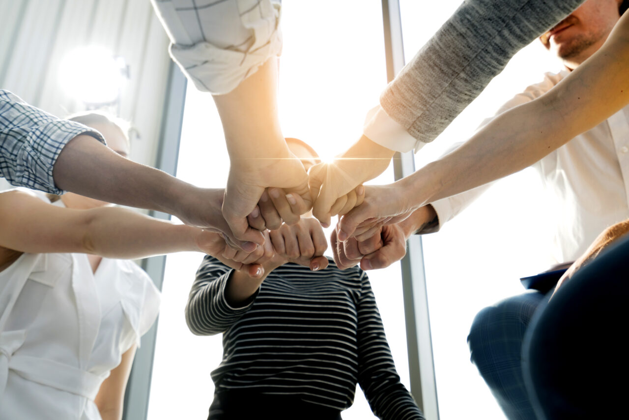 Eine Gruppe von Menschen steht im Kreis und hält die Hände im Zentrum des Bildes zusammen. Perspektive von schräg unten.