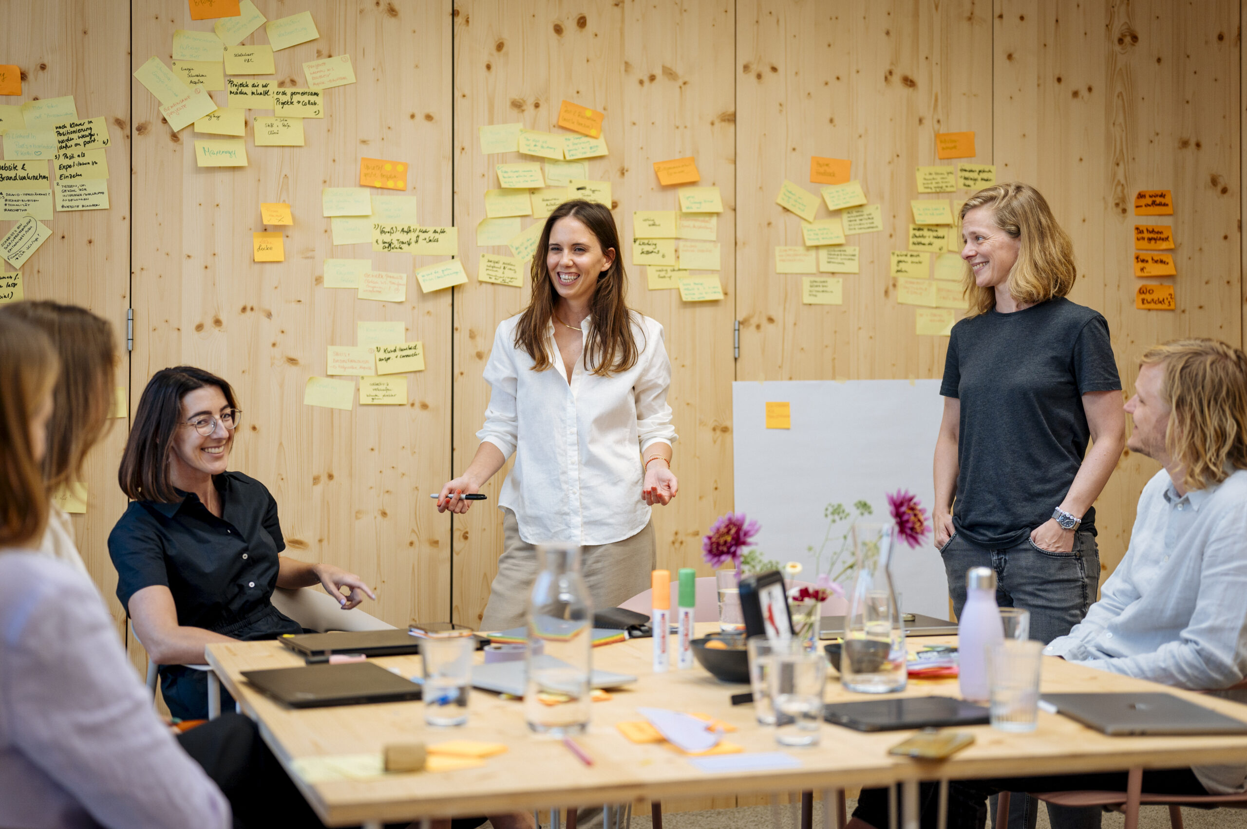 Eine Workshop-Szene: Junge Menschen vor einer Pinnwand mit Post-It's, davor ein Tisch mit Arbeitsutensilien und Gläsern