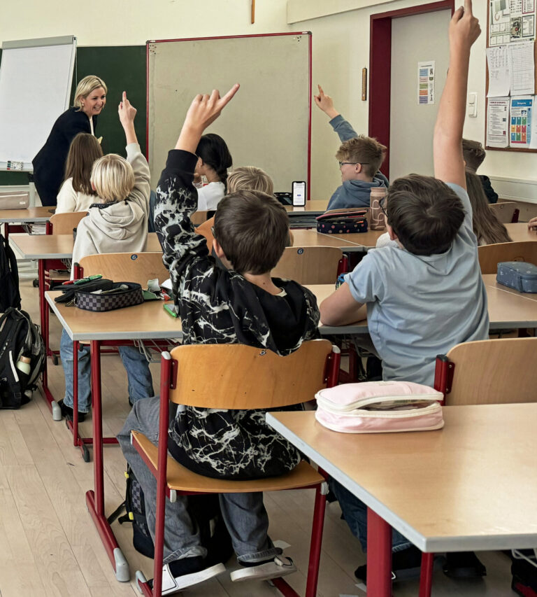 Blick ins Klassenzimmer einer Schule. Schüler:innen sitzen an ihren Schultischen und zeigen auf, im Hintergrund eine junge Lehrerin