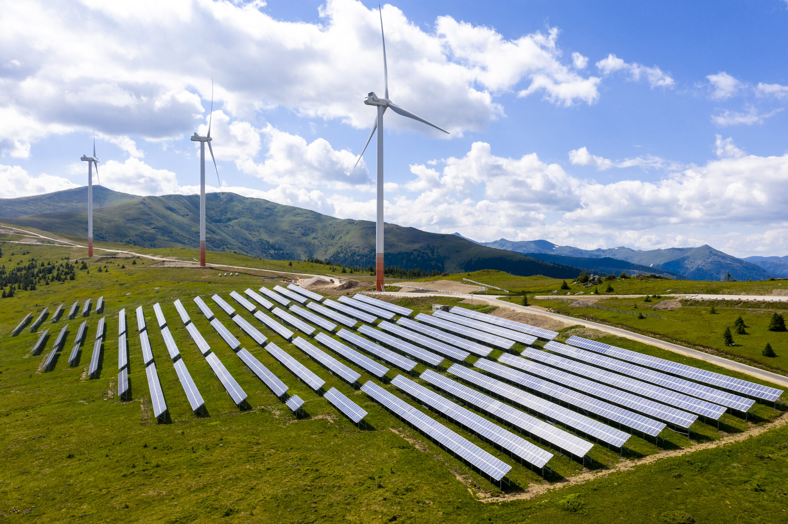 Drei Windräder auf einem Berggrat, davor eine große Anzahl von Solarpanelen auf einer Wiese. Im Hintergrund der Himmel und einige Wolken.